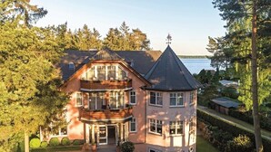 A large, multi-story house with a prominent turret and a balcony, surrounded by lush greenery and a body of water in the background.