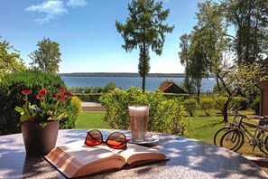 Un livre ouvert, une paire de lunettes de soleil et un verre de lait sur une table, avec vue sur un lac et un vélo en arrière-plan.