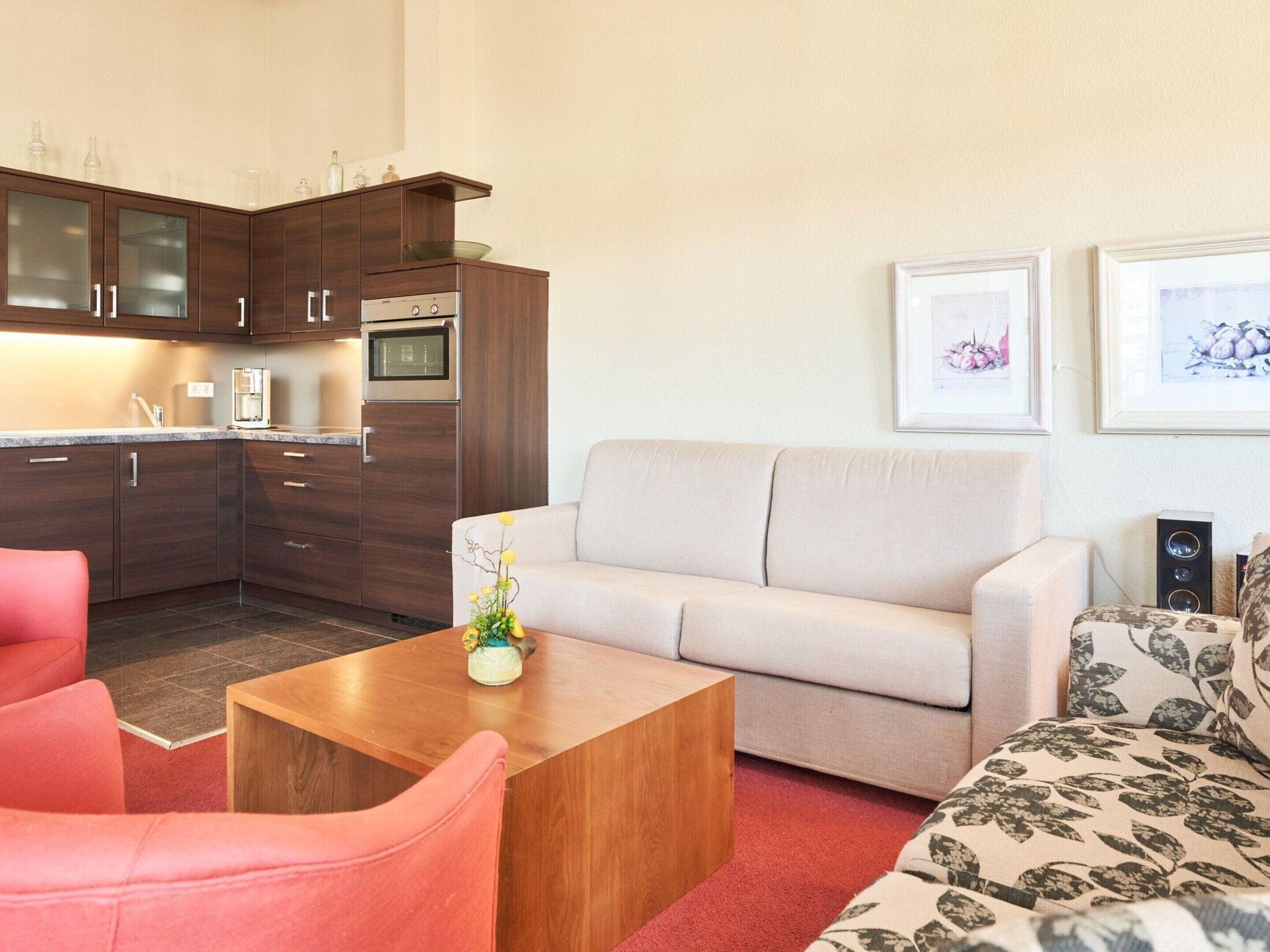 A modern kitchen with dark wood cabinets and a built-in oven. There is a beige sofa and a red armchair in the living area, with a wooden coffee table in between.