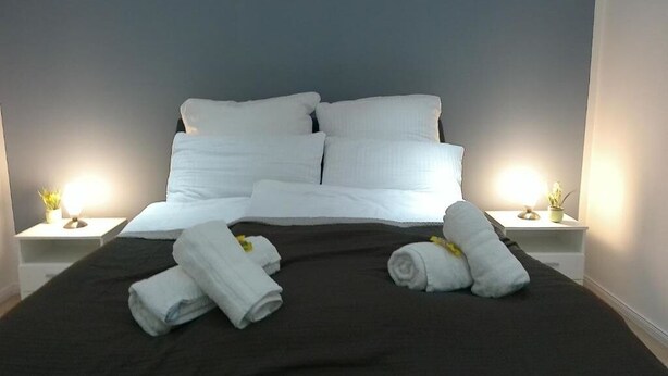 Ein ordentlich bezogenes Bett mit weißen Bettwäsche und Handtüchern, zwei Nachttische mit Lampen und eine dunkel gefärbte Tagesdecke.