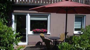 An outdoor patio with a table and two chairs, a red umbrella, and a window with pink flowers. - Garden-view Apartment in Heringhausen Lake City Centre (Diemelsee)