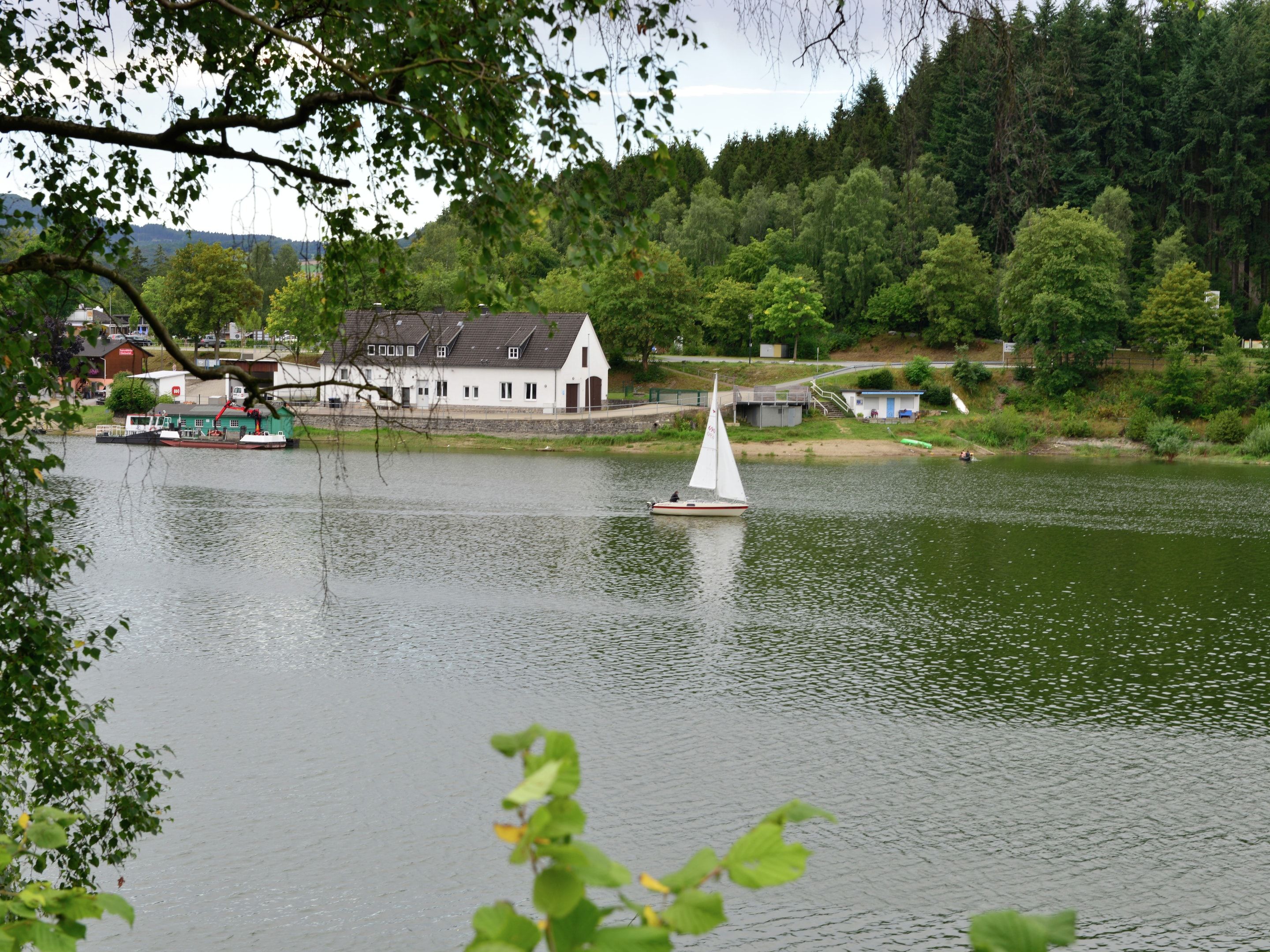 Un lago tranquilo con un velero, una casa blanca y un muelle con una grúa.