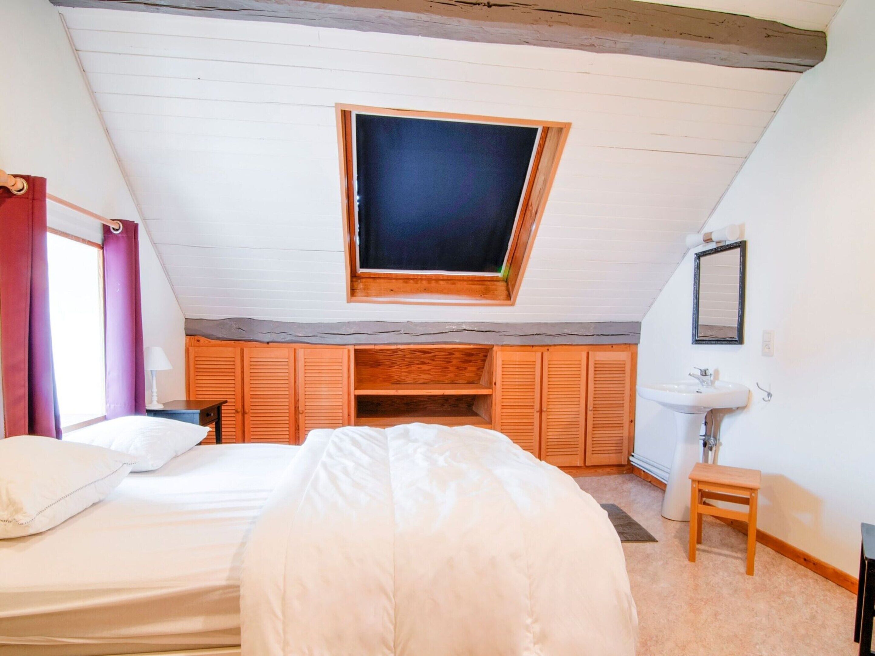 Ein Schlafzimmer mit schrägen Wänden, einem Oberlicht, einem Bett, einem Nachttisch, einem kleinen Holzschemel und einem Badezimmer mit Waschbecken und Spiegel.