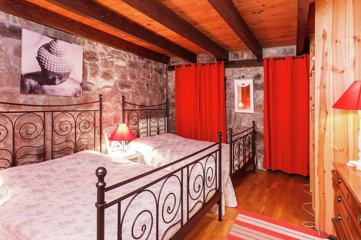 A bedroom with a stone wall, a bed with a headboard, a nightstand, a red lamp, and a wooden dresser.