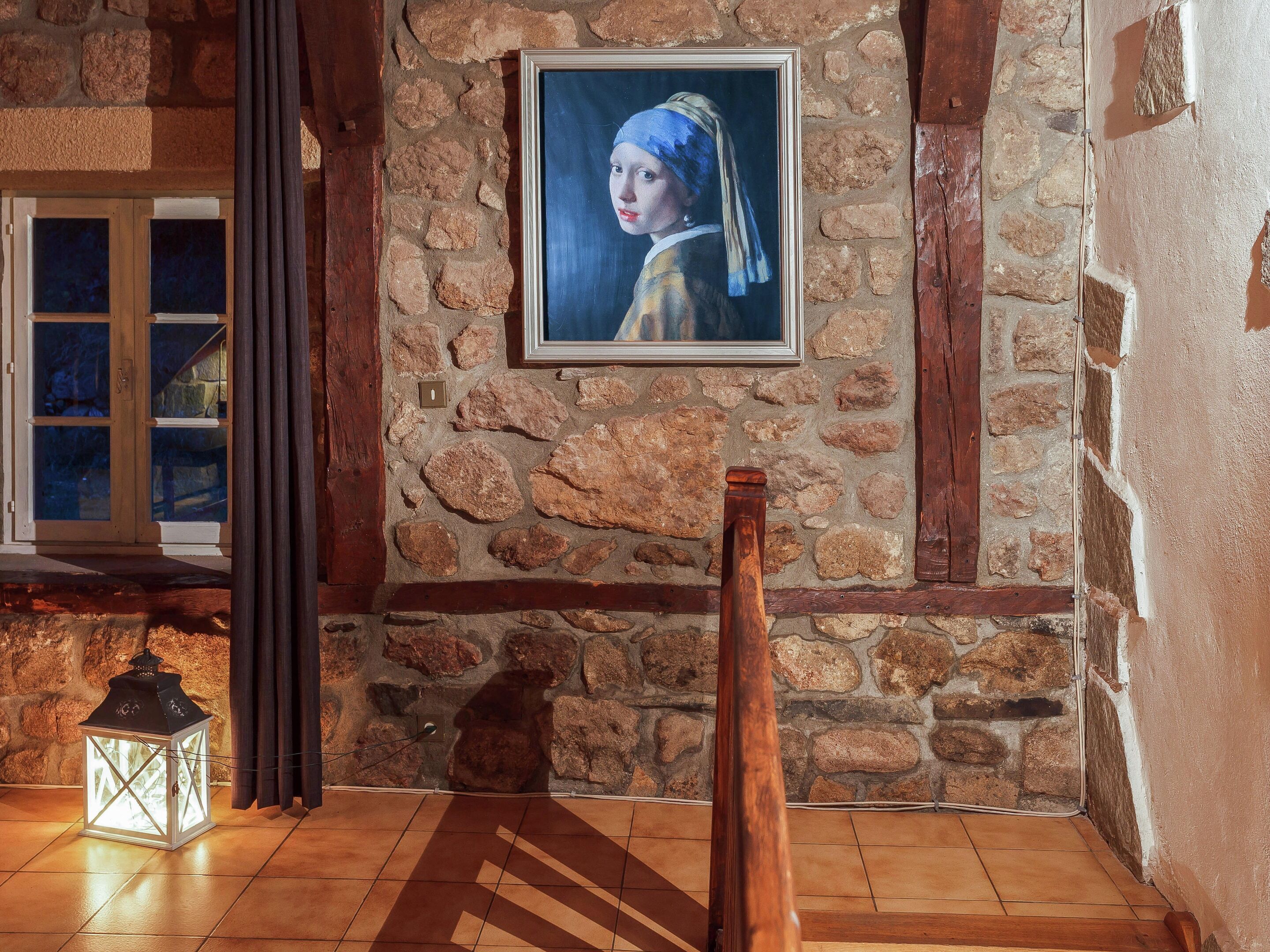 Quarto com paredes de pedra, piso de madeira, quadro emoldurado, lanterna e janela com cortinas.