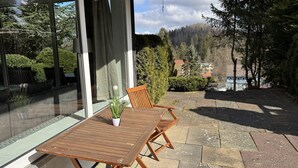 Une table et des chaises en bois sont placées à l’extérieur d’un bâtiment doté d’une porte en verre, le tout étant entouré de verdure et offrant une vue sur des maisons en arrière-plan.