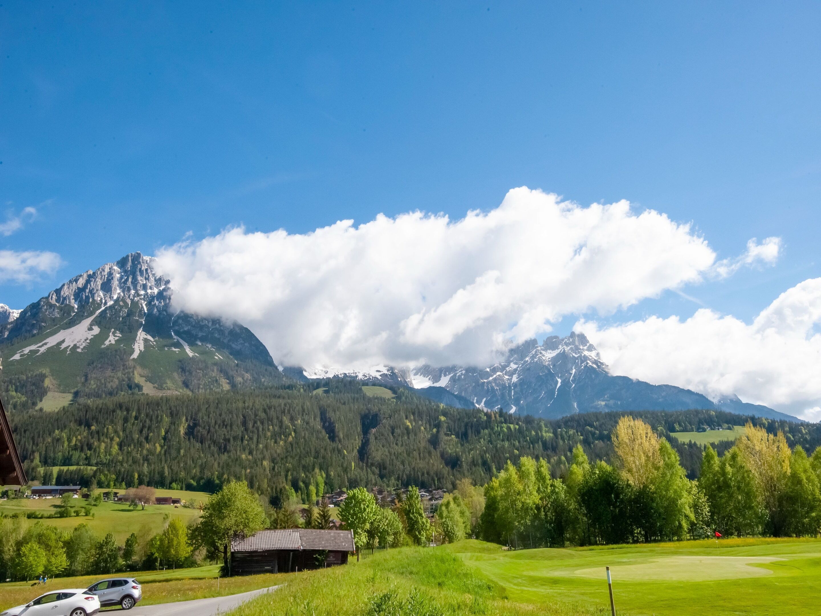 Un paysage de montagne avec un terrain de golf, une petite cabane en bois et quelques voitures garées à proximité.