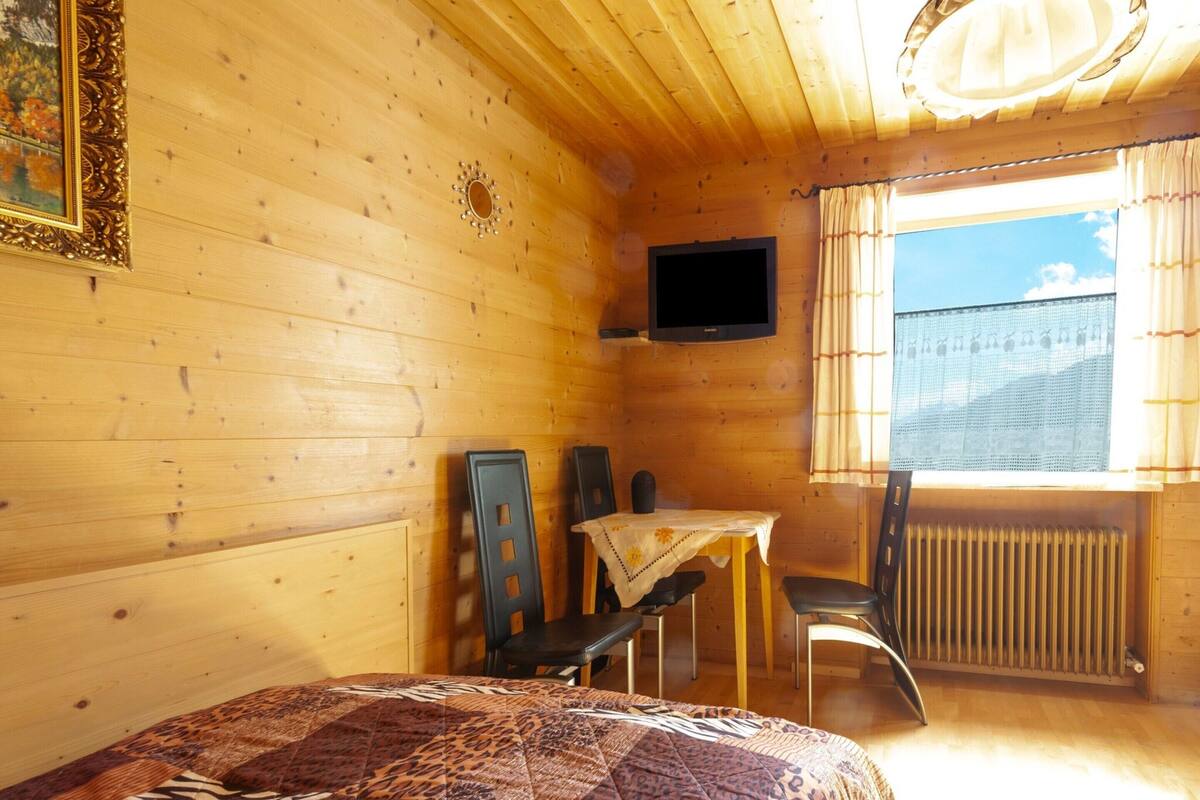 A cozy wooden cabin room with a bed, a table, two chairs, a TV, and a window with curtains.
