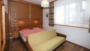 Ein Zimmer mit einem Bettgestell aus Holzlatten, einer Couch, einem Bett mit karierter Bettwäsche, einer Hängeleuchte und einem Fenster mit transparenten Vorhängen.