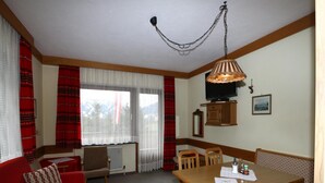 Ein gemütliches Zimmer mit einem roten Sofa, einem Holzesstisch und Stühlen. Ein Fernseher hängt an der Wand und es gibt ein Fenster mit Vorhängen.
