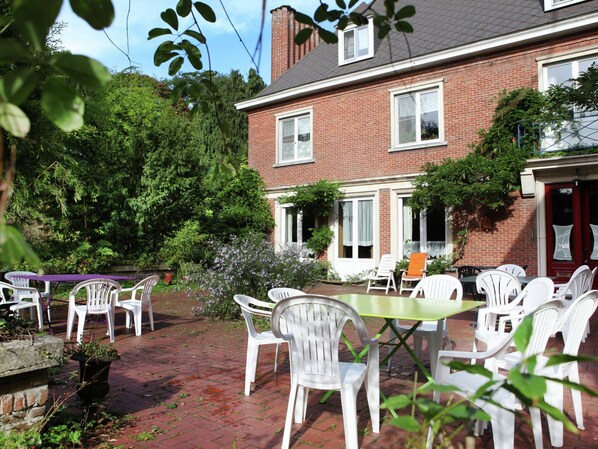 A patio area with white plastic chairs and tables, surrounded by greenery and a brick building.