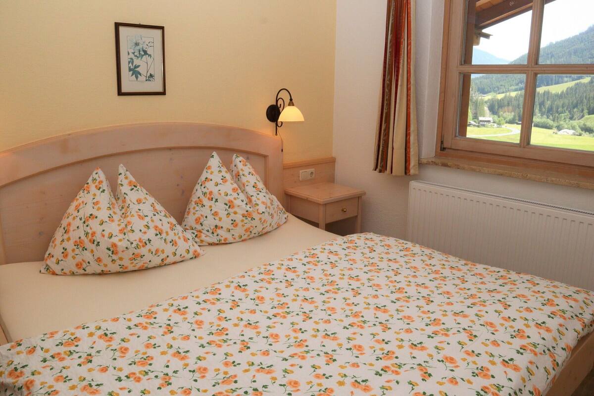 Ein Schlafzimmer mit einem Bett, einem Nachttisch, einer Lampe, einem Fenster mit Vorhängen und einem gerahmten Bild an der Wand.