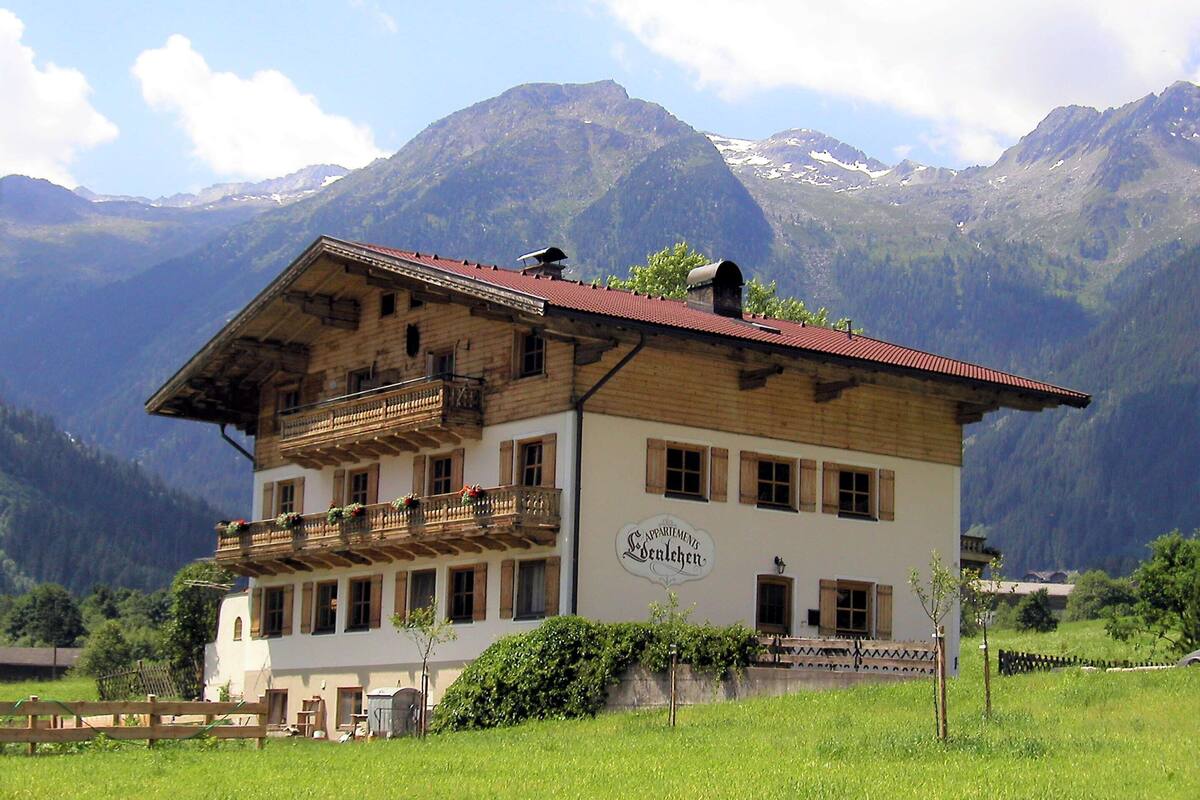 A traditional wooden building with a balcony, surrounded by a lush green field and mountains in the background.