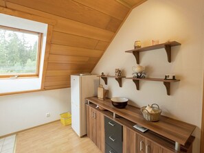 Eine Küche mit Holzschränken, einem Kühlschrank und einem Fenster mit Blick auf Bäume. Über der Arbeitsplatte befinden sich Regale mit Küchenutensilien.