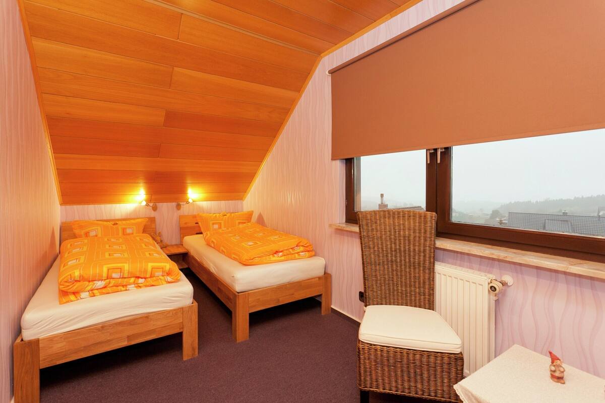 A bedroom with two wooden beds, a wicker chair, a small table, and a window with a view.