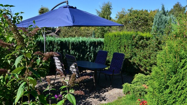 Ein Garten-Terrasse mit Tisch und Stühlen unter einem blauen Sonnenschirm, umgeben von üppigem Grün und Pflanzen.