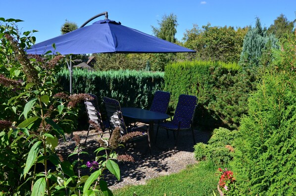Een terras in de tuin met een tafel en stoelen onder een blauwe parasol, omringd door weelderige begroeiing en planten.