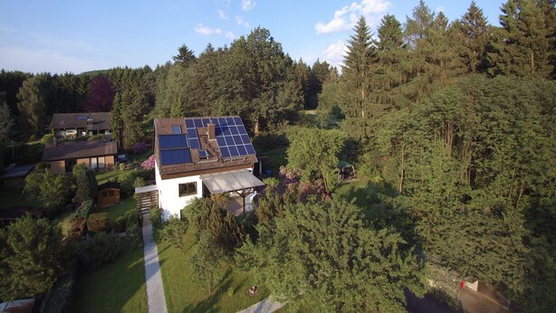 Une maison dotée d’un toit recouvert de panneaux solaires, entourée d’une forêt dense et accessible par une allée pavée.