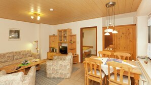 Ein gemütliches Wohnzimmer mit einer Couch, einem Couchtisch und einem Essbereich mit Tisch und Stühlen. Im Hintergrund befinden sich ein Fernseher und Holzschränke.