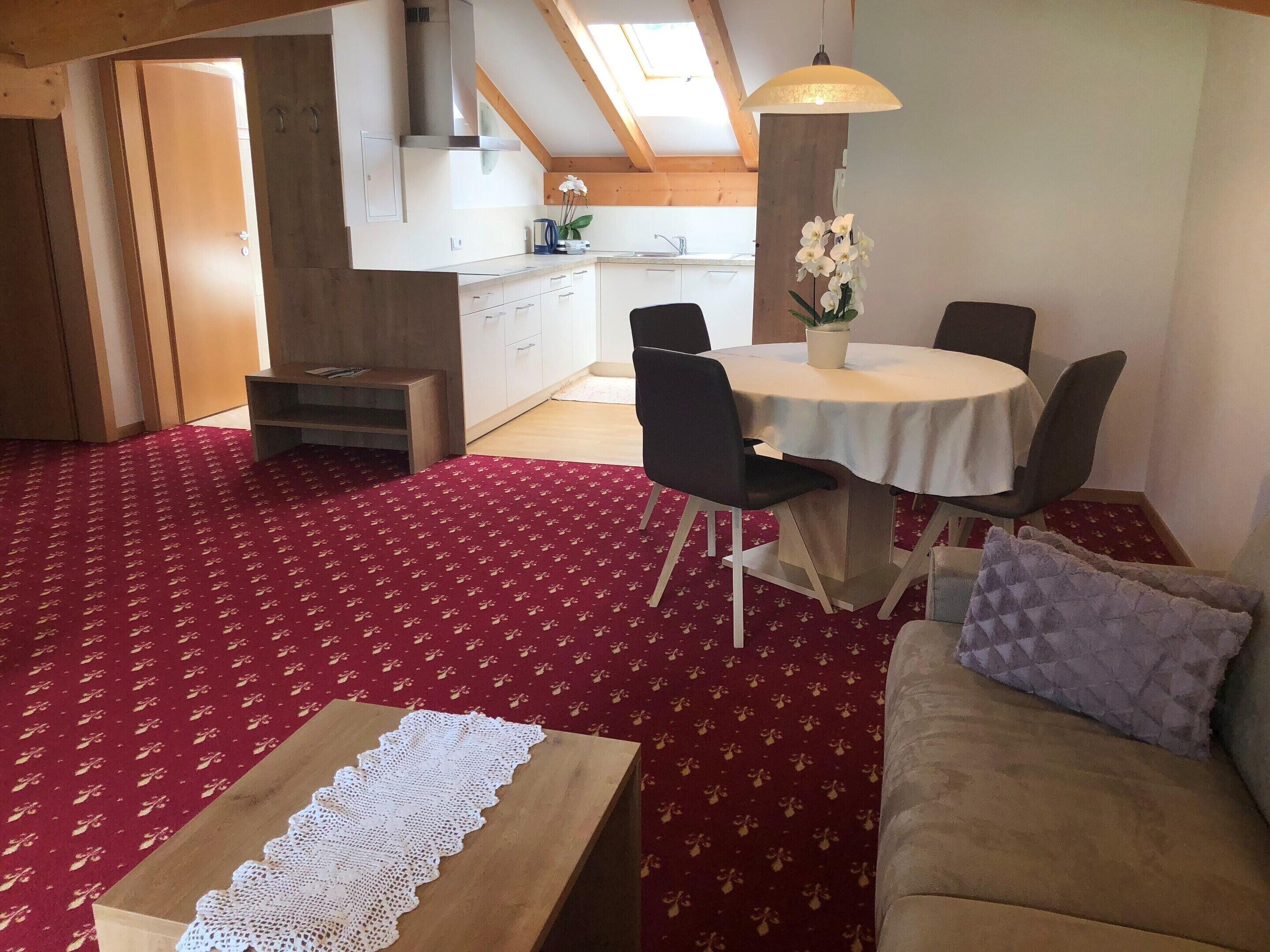 A cozy living room with a red patterned carpet, a wooden coffee table, a beige sofa with a pillow, and a dining area with a white table and chairs.