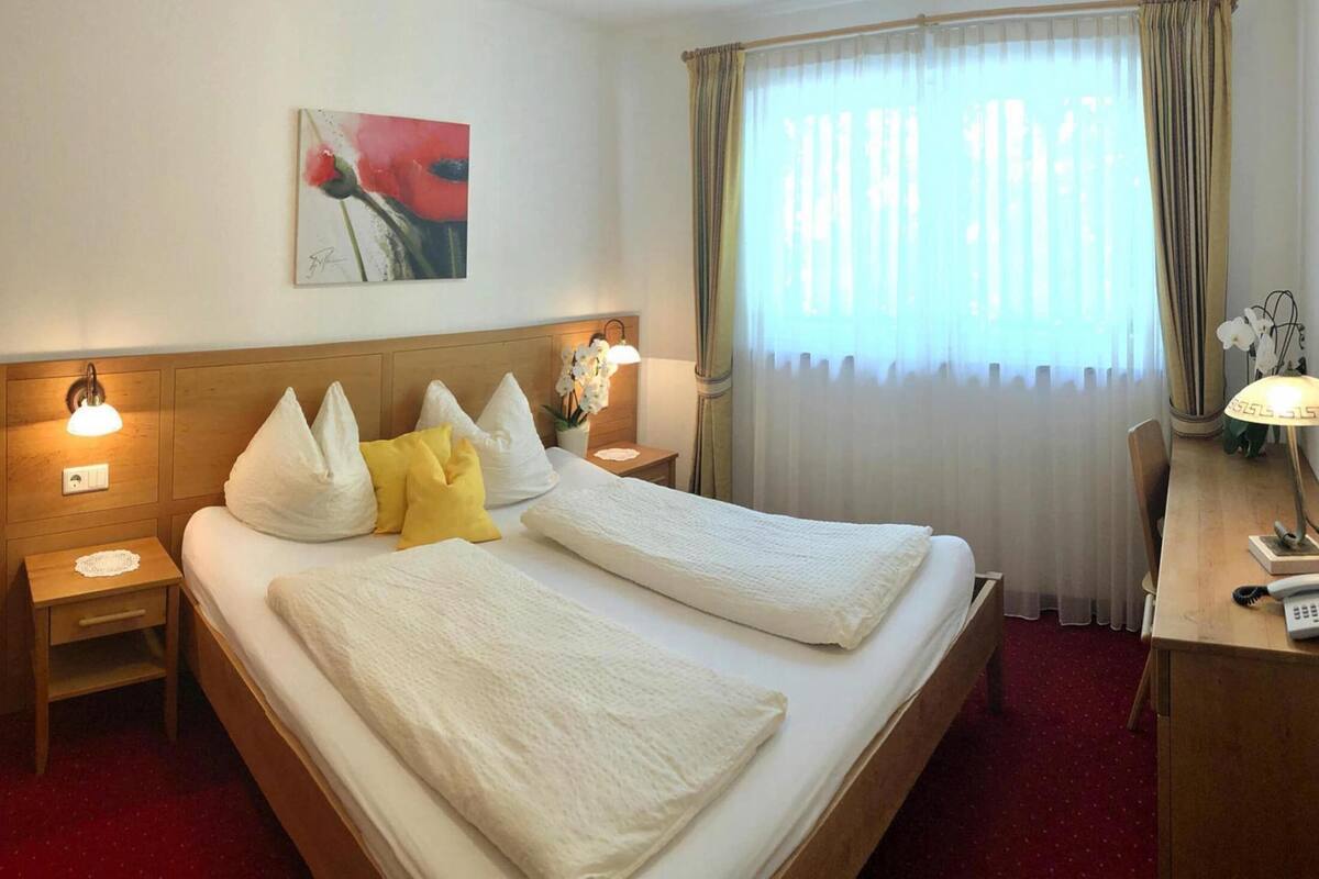 Ein Hotelzimmer mit einem Doppelbett, zwei Nachttischen mit Lampen, einem Schreibtisch mit Telefon und einem Fenster mit Vorhängen.