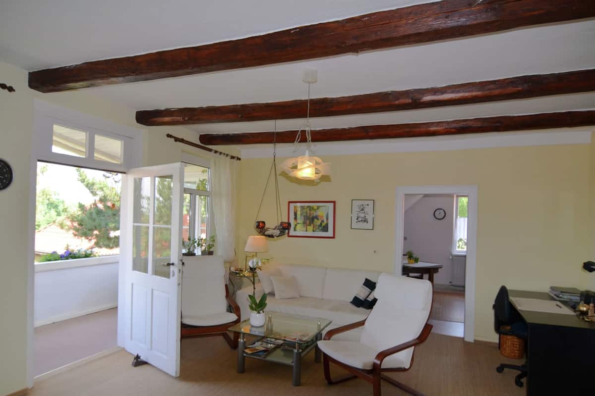 A living room with a white sofa, a glass coffee table, and a wooden ceiling beam. There is a door leading to another room and a window with curtains.