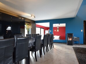 A modern dining area with a long table and chairs, a kitchen in the background, and a fireplace. - Modern Cottage in Vaux-sur-sure With Sauna (Vaux-sur-Sure)