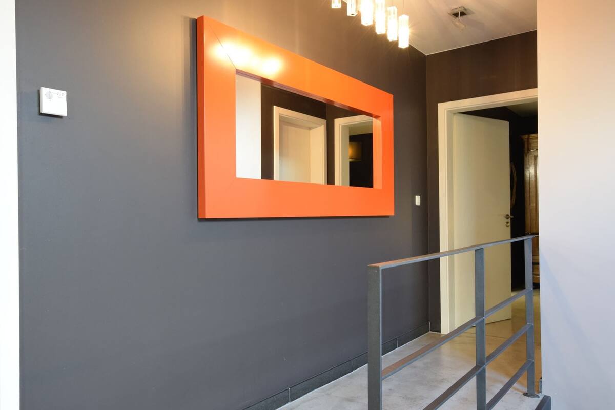 A hallway with a dark gray wall, a bright orange-framed mirror, and a metal railing.