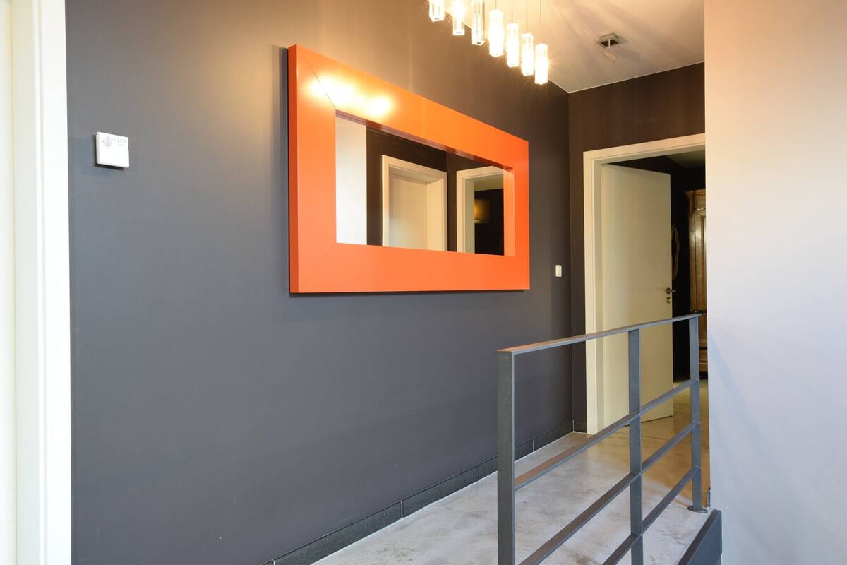 A hallway with a grey wall, a bright orange mirror, and a metal railing.