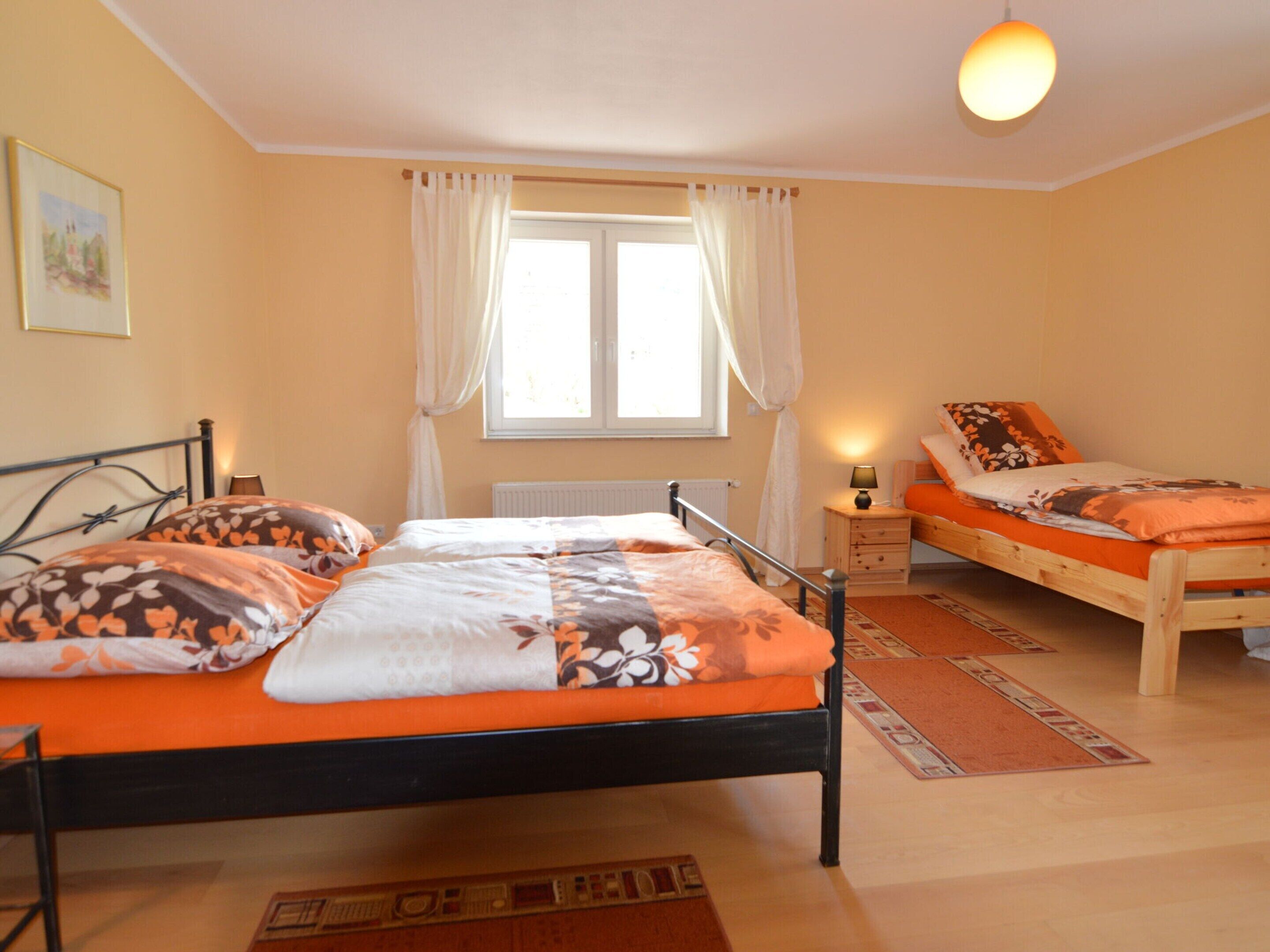 A bedroom with two beds, a window with curtains, a bedside table with a lamp, and a radiator.
