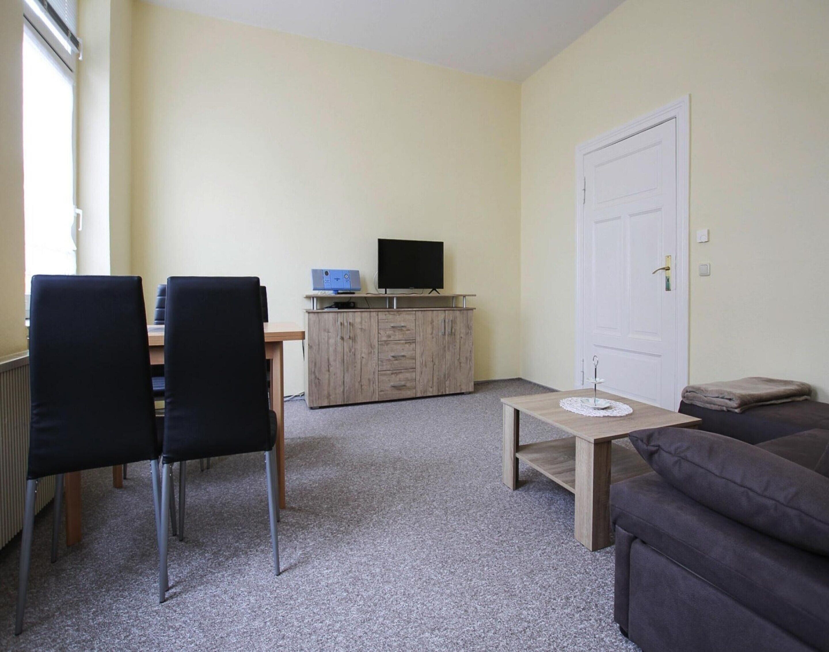 A small, furnished room with a desk, two chairs, a television, a sofa, and a door.