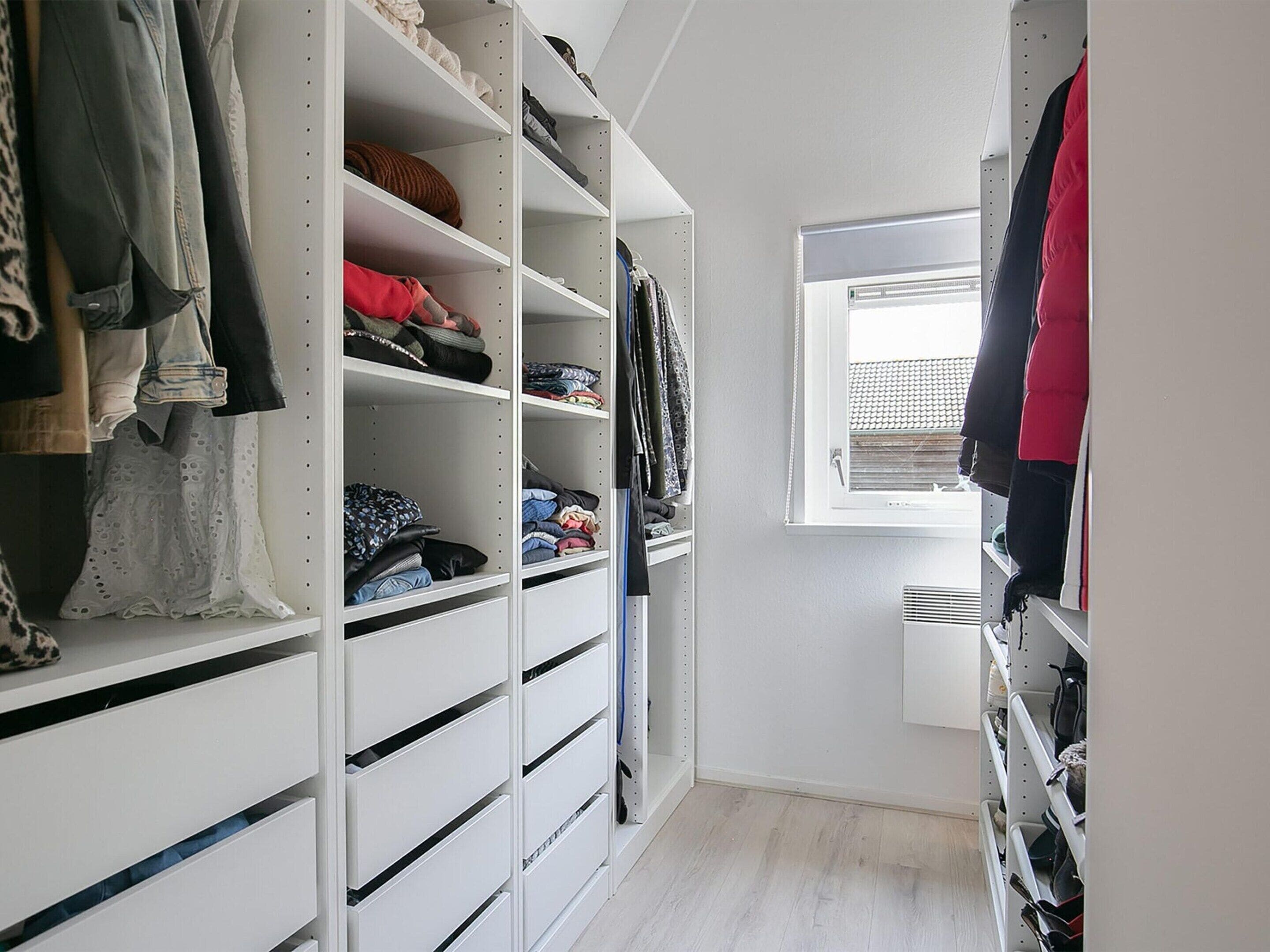 A walk-in closet with multiple shelves and drawers for storage, a window with blinds, and a wooden floor.