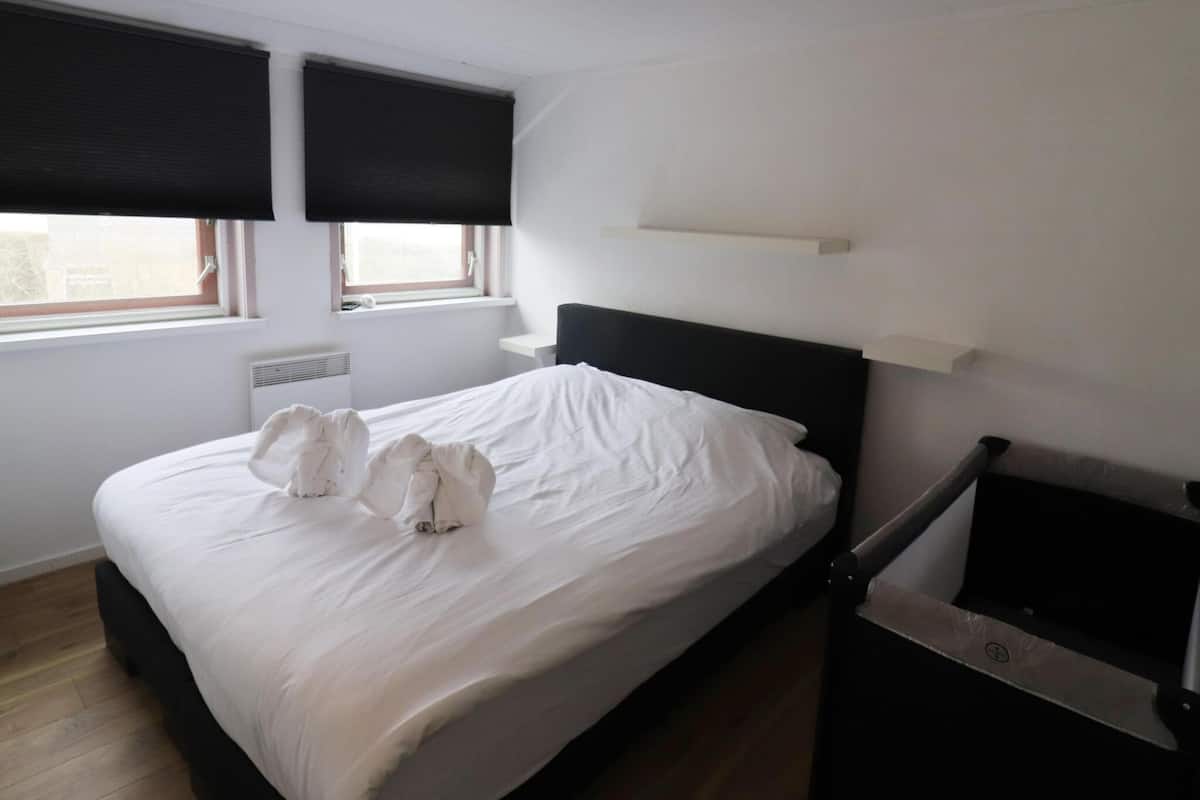 A neatly made bed with white linens and two towels placed on the bed. There is a black headboard, a small bedside table with a lamp, and a window with blinds.