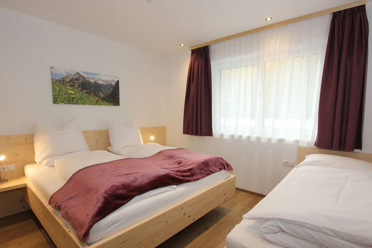 Une chambre avec un lit en bois, une literie blanche, une table de chevet avec une lampe, une fenêtre avec des rideaux et un tableau représentant un paysage de montagne accroché au mur.