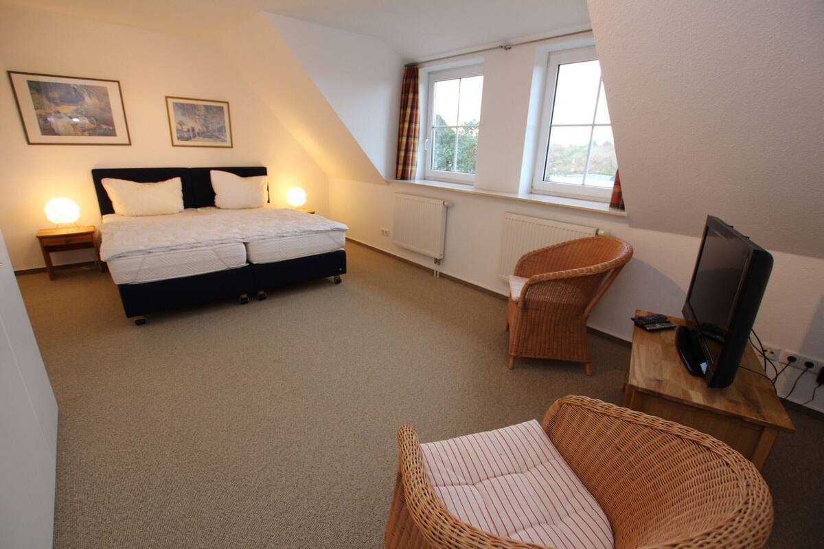 Een slaapkamer met een bed, twee bedlampjes, een televisie op een houten standaard en een rieten stoel.