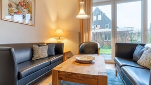 A modern living room with a black leather sofa, a wooden coffee table, and a large window with a view of a house and garden. - Terraced House, St. Peter - Ording (St. Peter-Ording)