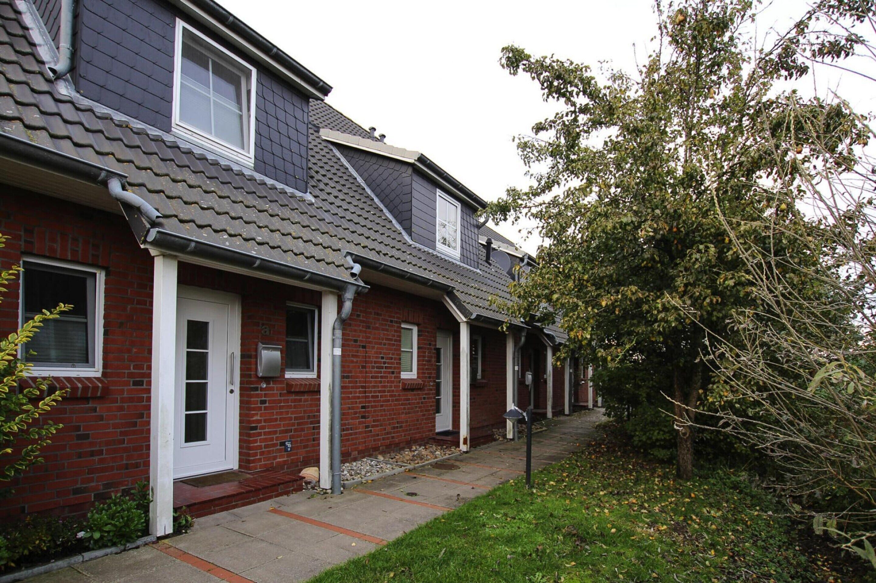 A row of brick townhouses with a grey tiled roof, a white door, and a small front yard with a tree.