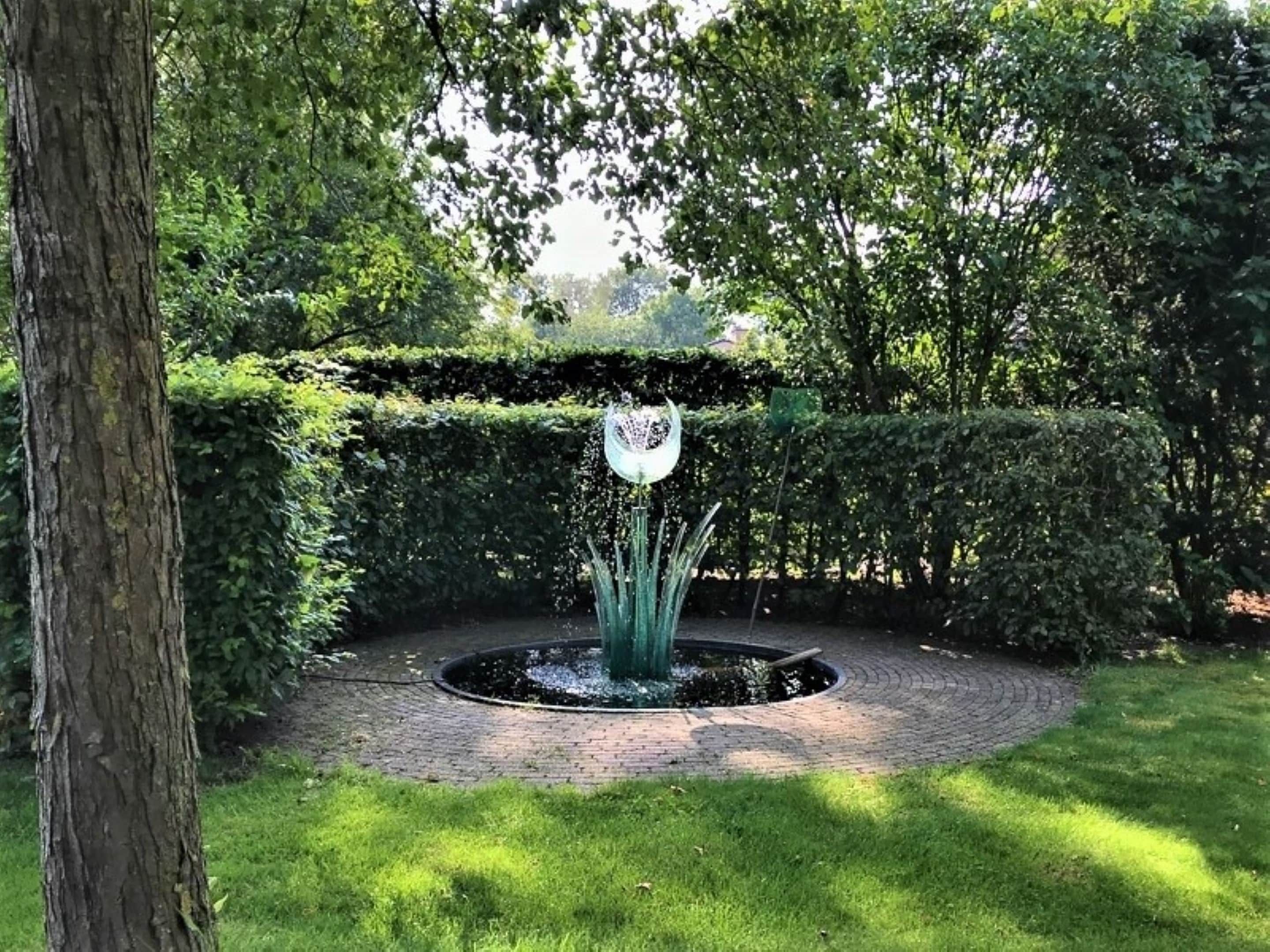 A circular brick pathway with a central water feature surrounded by well-maintained hedges and trees.
