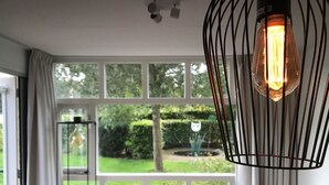 A room with a large window, a hanging light fixture, and a view of a garden. - Homely Apartment in Noordwolde With Balcony (Noordwolde)