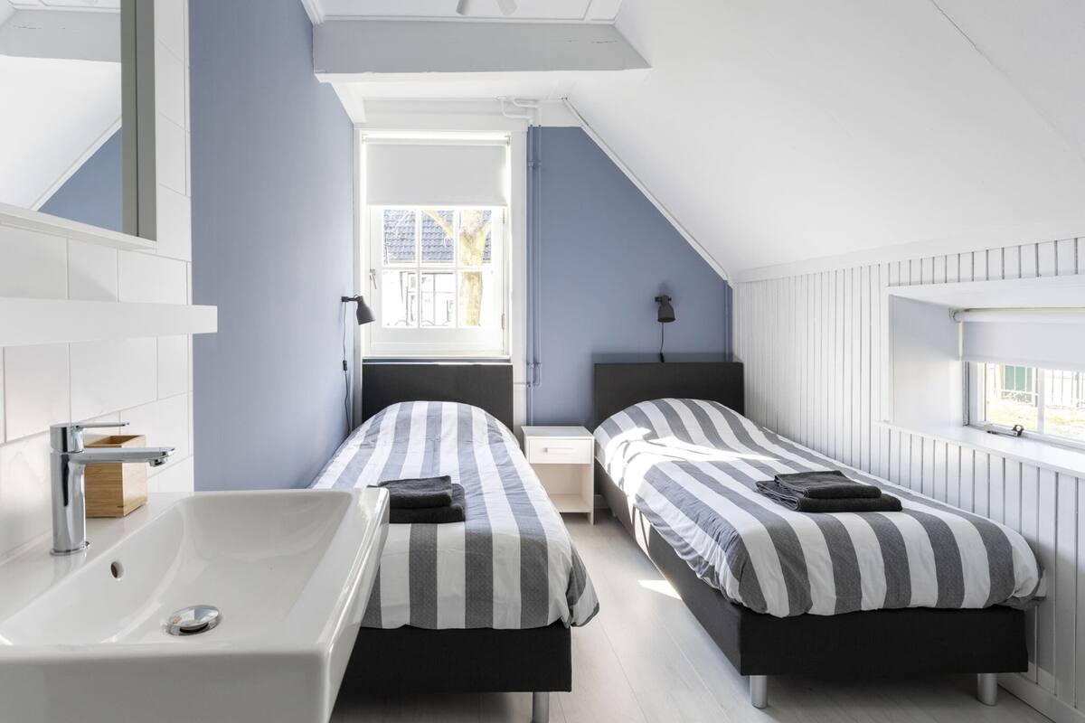 Een slaapkamer met twee bedden, een groot raam, een dakvenster, een witte badkuip en een houten kast.