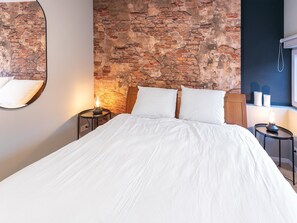 A bedroom with a brick wall, a bed with white bedding, two bedside tables with lamps, and a mirror on the wall.