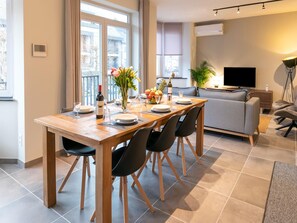 A modern living room with a dining area, a grey sofa, a flat-screen TV, and a large window with curtains. - Duplex Apartment in the Shopping Street of Maasmechelen (Maasmechelen)