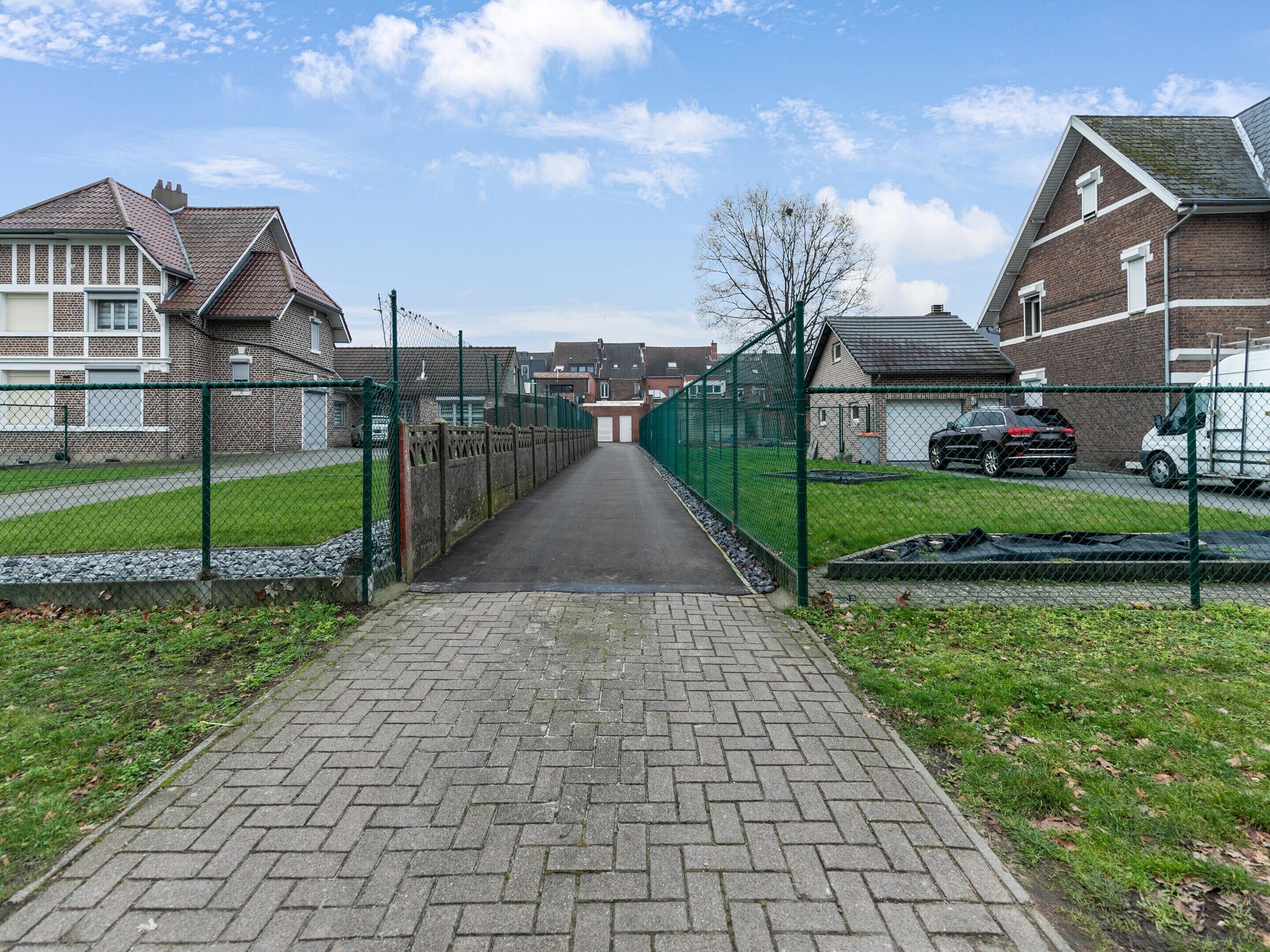 A paved pathway leading to a residential area with houses on either side, a green fence, and a clear sky.