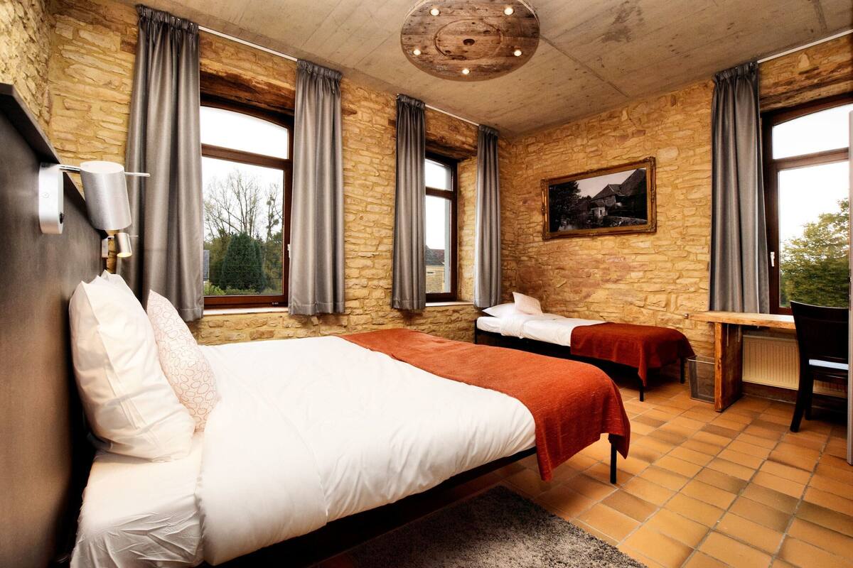 A room with a large bed, a smaller bed, a desk, and a chair. There are windows with curtains, a painting on the wall, and a stone wall.