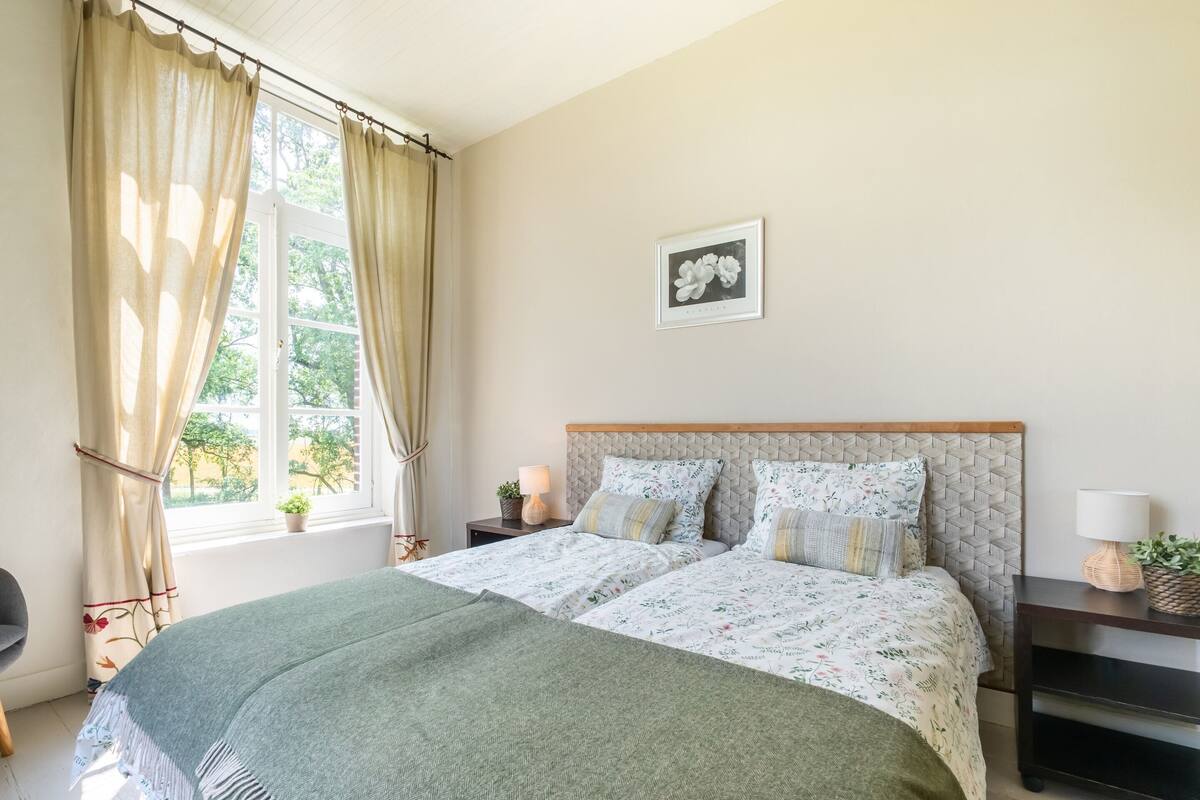 A bedroom with a bed, two bedside tables, a window with curtains, a wall-mounted picture, and a chair.