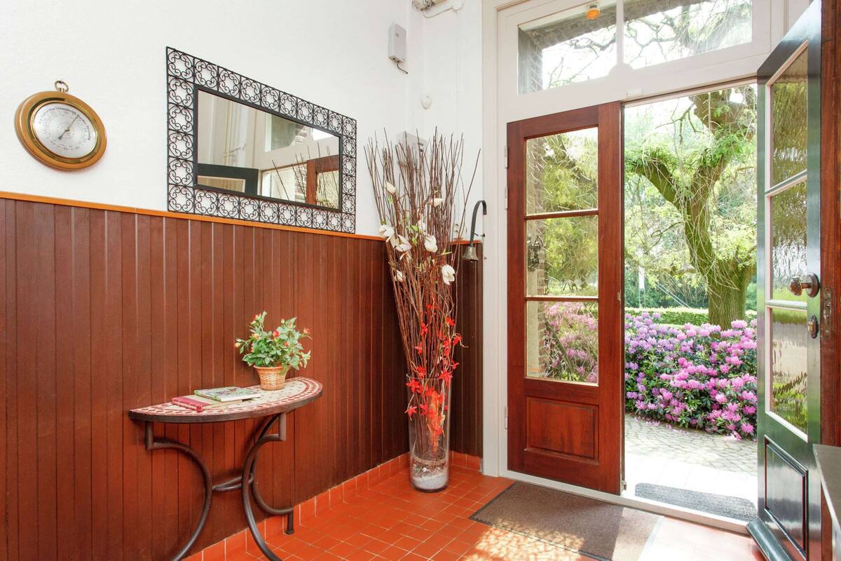 A wooden panel wall, a round table with a potted plant, a tall vase with branches, and a door leading to a garden.