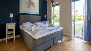 A bedroom with a bed, a nightstand, a lamp, a painting, and a sliding glass door leading to a garden.