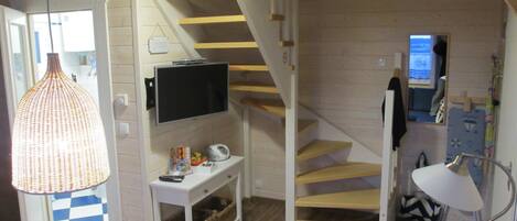 Un espacio habitable compacto con una escalera, un televisor montado en la pared, una pequeña mesa con una cesta y una lámpara colgante.
