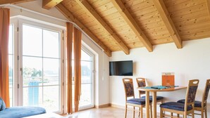 Ein Zimmer mit Holzboden, einem an der Wand befestigten Fernseher, einem Tisch mit Stühlen und einem großen Fenster mit Vorhängen.