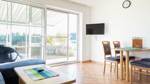 Ein Zimmer mit einer Couch, einem kleinen Tisch mit Unterlage, einem an der Wand befestigten Fernseher und einem Essbereich mit Stühlen und Tisch.
